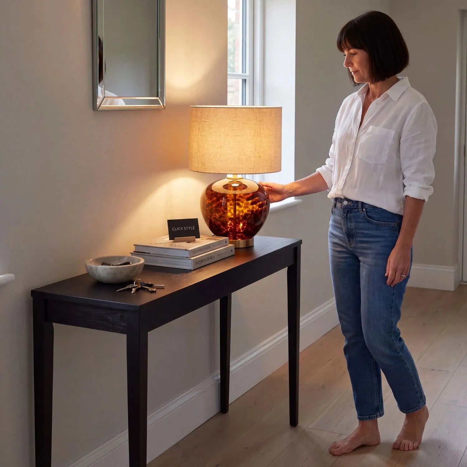 Amber & Brown Tortoiseshell Glass Table Lamp with Natural Linen Shade styled on a dark wood console table in a contemporary hallway with a woman switching on the lamp 44x30x30cm | Click Style