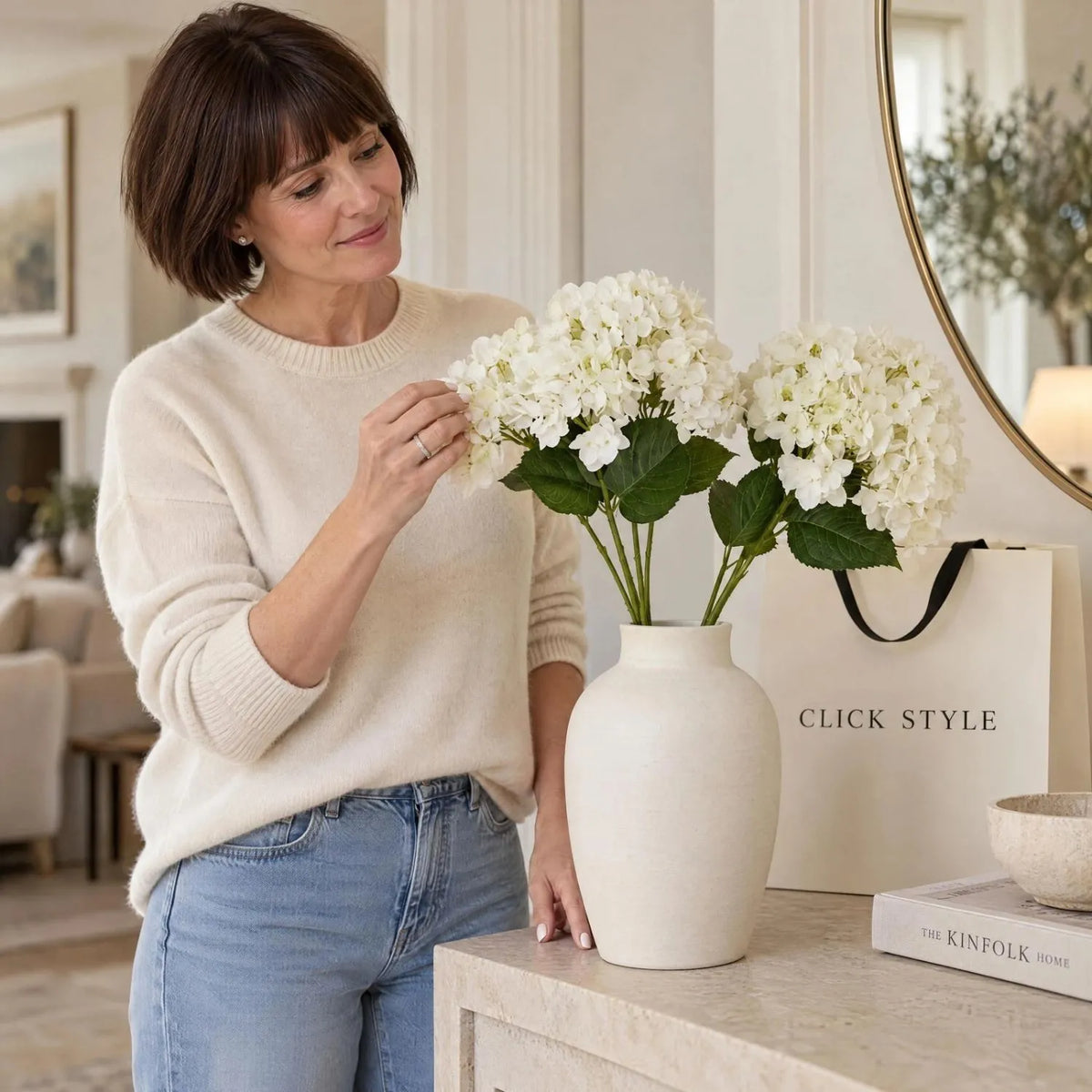 Artificial Creamy White Multi-Bloom Hydrangea Stem with Realistic Green Leaves – Lifelike Faux White Hydrangea Flower 47x23x23cm – Woman in cream cashmere jumper arranging white faux hydrangeas in a ceramic vase beside CLICK STYLE shopping bag | Click Style