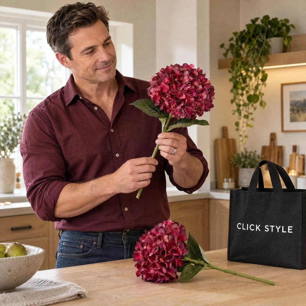 Artificial Deep Ruby Red & Burgundy Hydrangea Stem with Realistic Green Leaves – Lifelike Faux Red Hydrangea Flower 42x18x18cm – Man Wearing a Burgundy Shirt Examining a Ruby Red Artificial Hydrangea Stem with a Second Stem on a Pale Oak Kitchen Worktop and Black CLICK STYLE Branded Tote Bag | Click Style