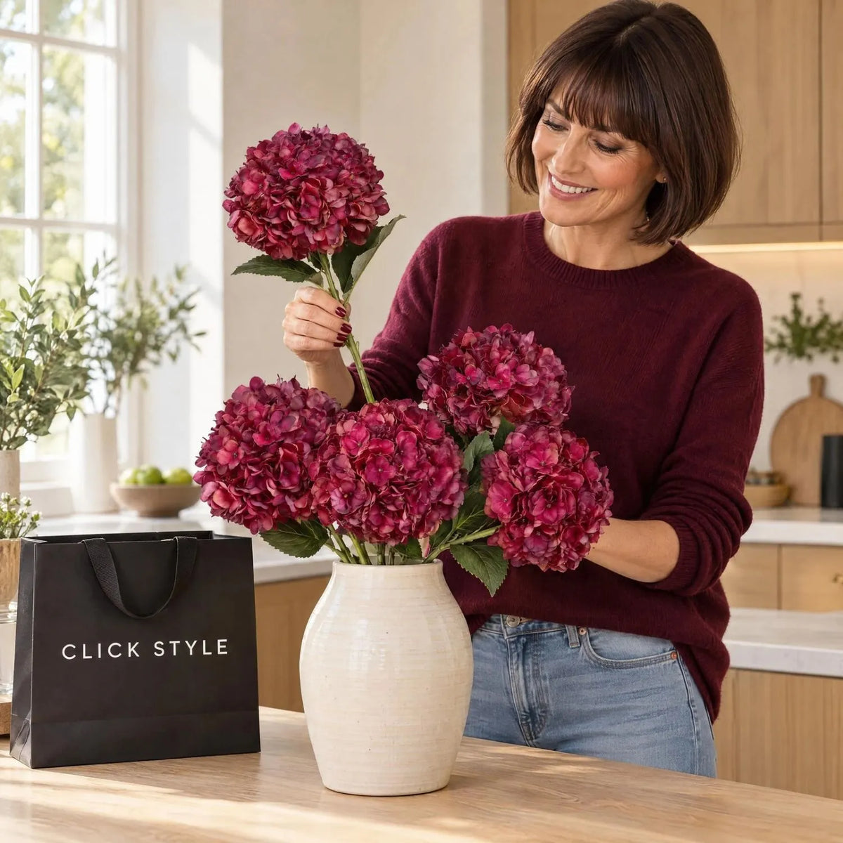 Artificial Deep Ruby Red & Burgundy Hydrangea Stem with Realistic Green Leaves – Lifelike Faux Red Hydrangea Flower 42x18x18cm – Woman Wearing a Burgundy Cashmere Jumper Arranging Five Artificial Hydrangea Stems in a Cream Ceramic Vase on a Kitchen Island with Black CLICK STYLE Branded Bag | Click Style