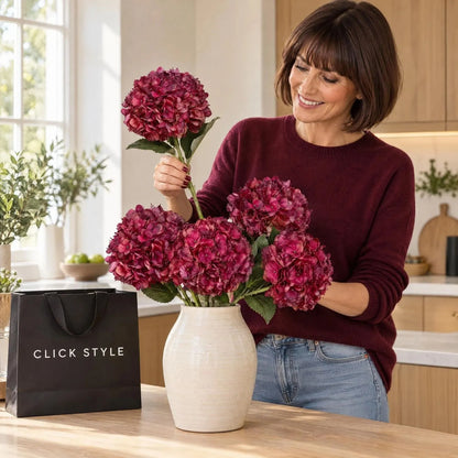 Artificial Deep Ruby Red & Burgundy Hydrangea Stem with Realistic Green Leaves – Lifelike Faux Red Hydrangea Flower 42x18x18cm – Woman Wearing a Burgundy Cashmere Jumper Arranging Five Artificial Hydrangea Stems in a Cream Ceramic Vase on a Kitchen Island with Black CLICK STYLE Branded Bag | Click Style