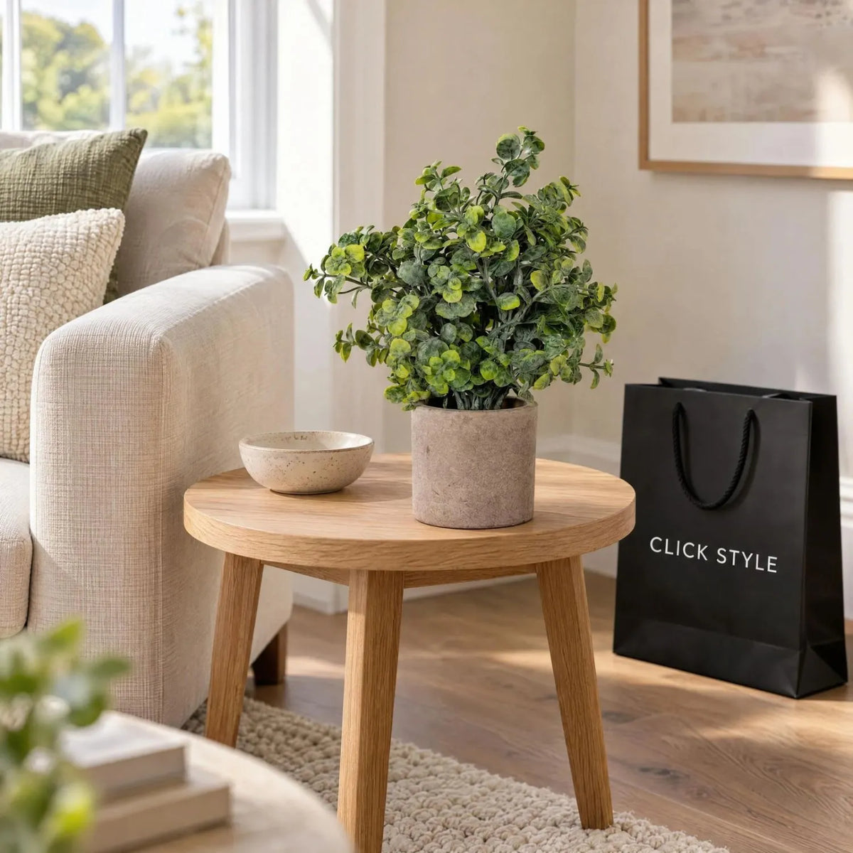 Artificial Green Eucalyptus Plant with Realistic Rounded Leaves in Rustic Stone-Effect Pot – Small Faux Potted Eucalyptus 23x19x19cm displayed on a round oak side table beside a cream sofa and ceramic bowl with black CLICK STYLE shopping bag | Click Style