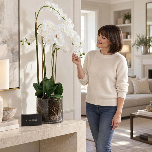 Artificial Large White Orchid with Cascading Real-Touch Blooms & Realistic Exposed Roots in Clear Glass Pot – Tall Faux Orchid Arrangement 126x57x57cm displayed on a stone console table beside a woman in a cream jumper showing the realistic white flowers | Click Style