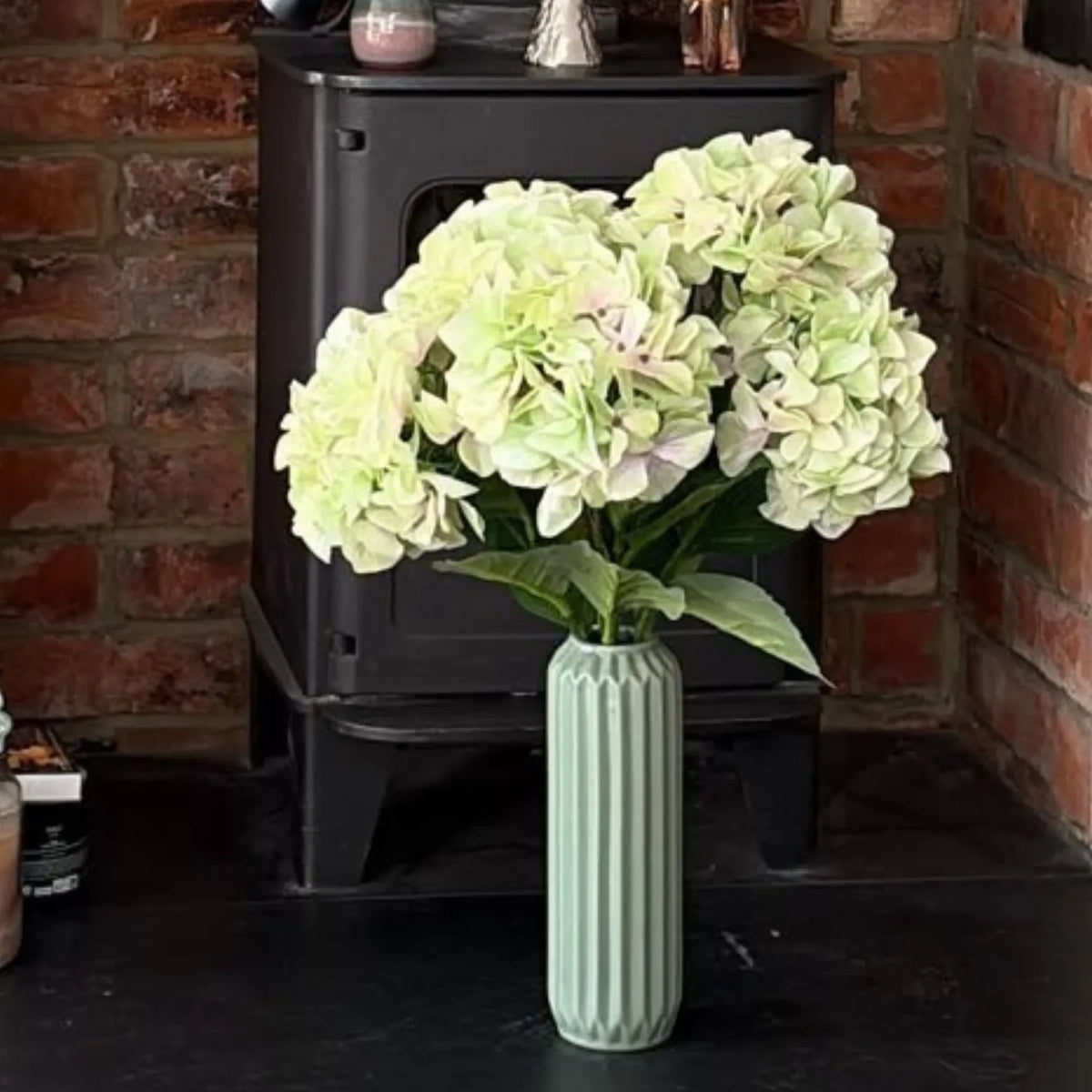 Artificial Pale Green Multi-Bloom Hydrangea Bouquet Stem with Realistic Foliage – Large Lifelike Faux Light Lime Green Hydrangea 58cm – Customer Image Showing One Stem in a Pale Green Ribbed Vase Beside a Wood-Burning Stove | Click Style