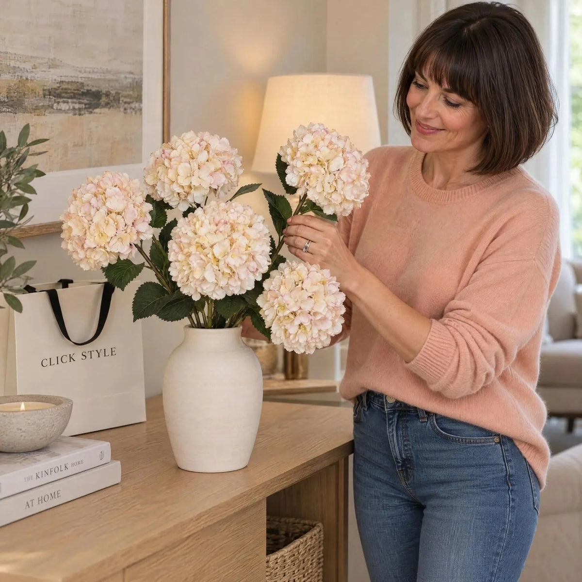 Artificial Peach Blush & Cream Hydrangea Stem with Realistic Green Leaves – Lifelike Faux Pink Hydrangea Flower 42x18x18cm shown as five individual stems being arranged in a ceramic vase by a woman wearing a peach cashmere jumper | Click Style