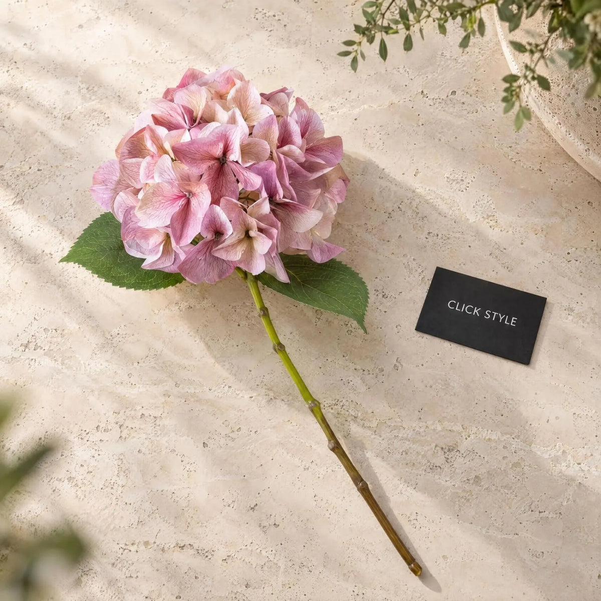 Artificial Pink Hydrangea Stem with Multi-Tonal Petals & Realistic Green Leaves – Lifelike Faux Pink Hydrangea Flower 50x30x30cm displayed individually on a natural travertine surface, showing its rounded pink and mauve flower head, delicate petal veining, two serrated green leaves and long green-brown stem | Click Style