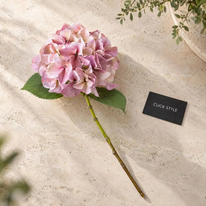Artificial Pink Hydrangea Stem with Multi-Tonal Petals & Realistic Green Leaves – Lifelike Faux Pink Hydrangea Flower 50x30x30cm displayed individually on a natural travertine surface, showing its rounded pink and mauve flower head, delicate petal veining, two serrated green leaves and long green-brown stem | Click Style