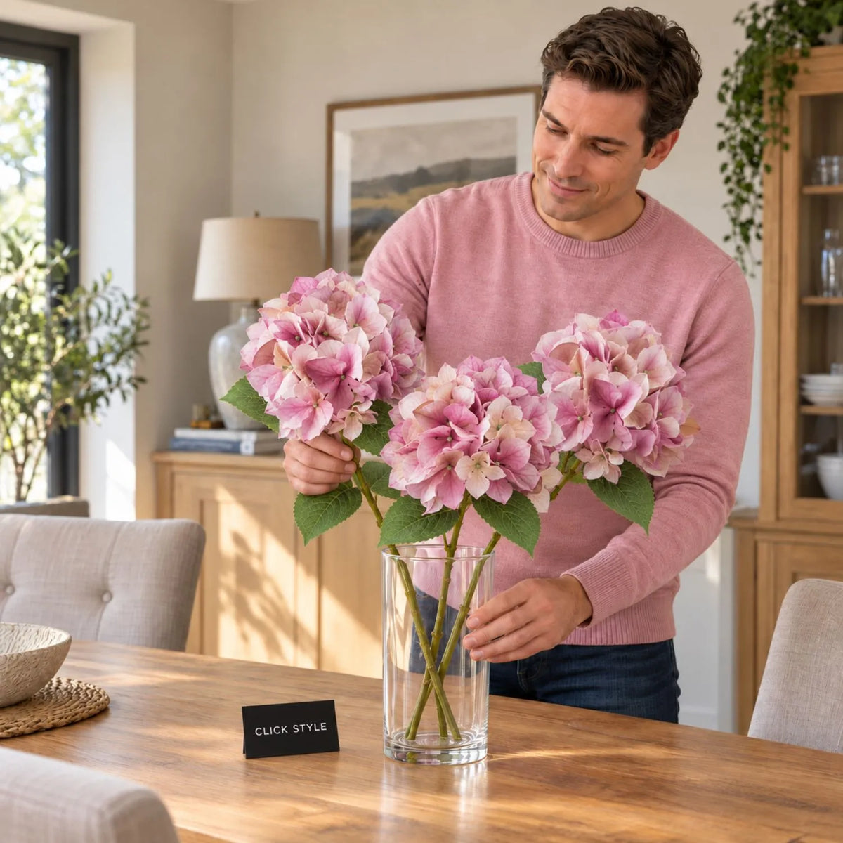 Artificial Pink Hydrangea Stem with Multi-Tonal Petals & Realistic Green Leaves – Lifelike Faux Pink Hydrangea Flower 50x30x30cm shown as three individual stems being arranged in a clear glass vase on an oak dining table by a man wearing a pink jumper, featuring layered pink, mauve and cream petals, delicate veining and serrated green leaves | Click Style