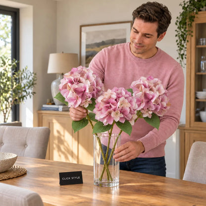 Artificial Pink Hydrangea Stem with Multi-Tonal Petals & Realistic Green Leaves – Lifelike Faux Pink Hydrangea Flower 50x30x30cm shown as three individual stems being arranged in a clear glass vase on an oak dining table by a man wearing a pink jumper, featuring layered pink, mauve and cream petals, delicate veining and serrated green leaves | Click Style