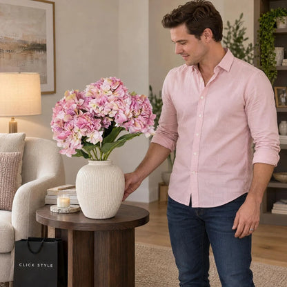 Artificial Pink Multi-Bloom Hydrangea Bouquet Stem with Realistic Green Leaves – Lifelike Faux Pink Hydrangea Flower Arrangement 58x47x47cm displayed in an ivory ceramic vase on dark oak side table beside man in pink shirt | Click Style