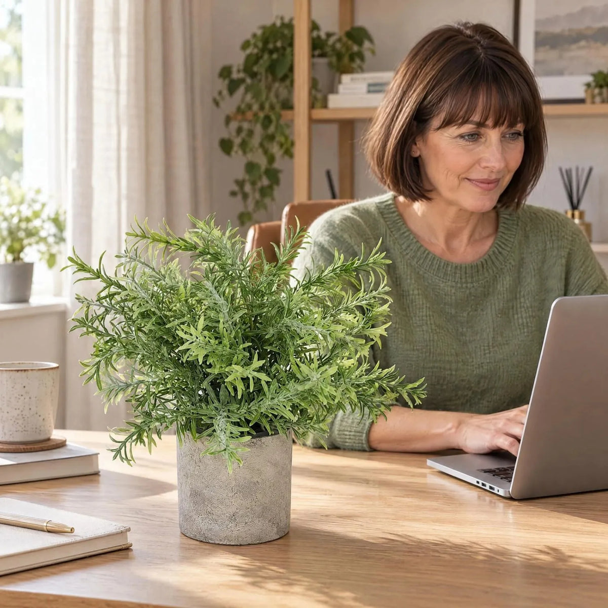 Artificial Rosemary Plant in Grey Stone-Effect Pot with Realistic Green Foliage – Lifelike Faux Potted Herb 24x24x24cm displayed on a light oak home office desk beside a woman in a sage-green jumper working on her laptop | Click Style