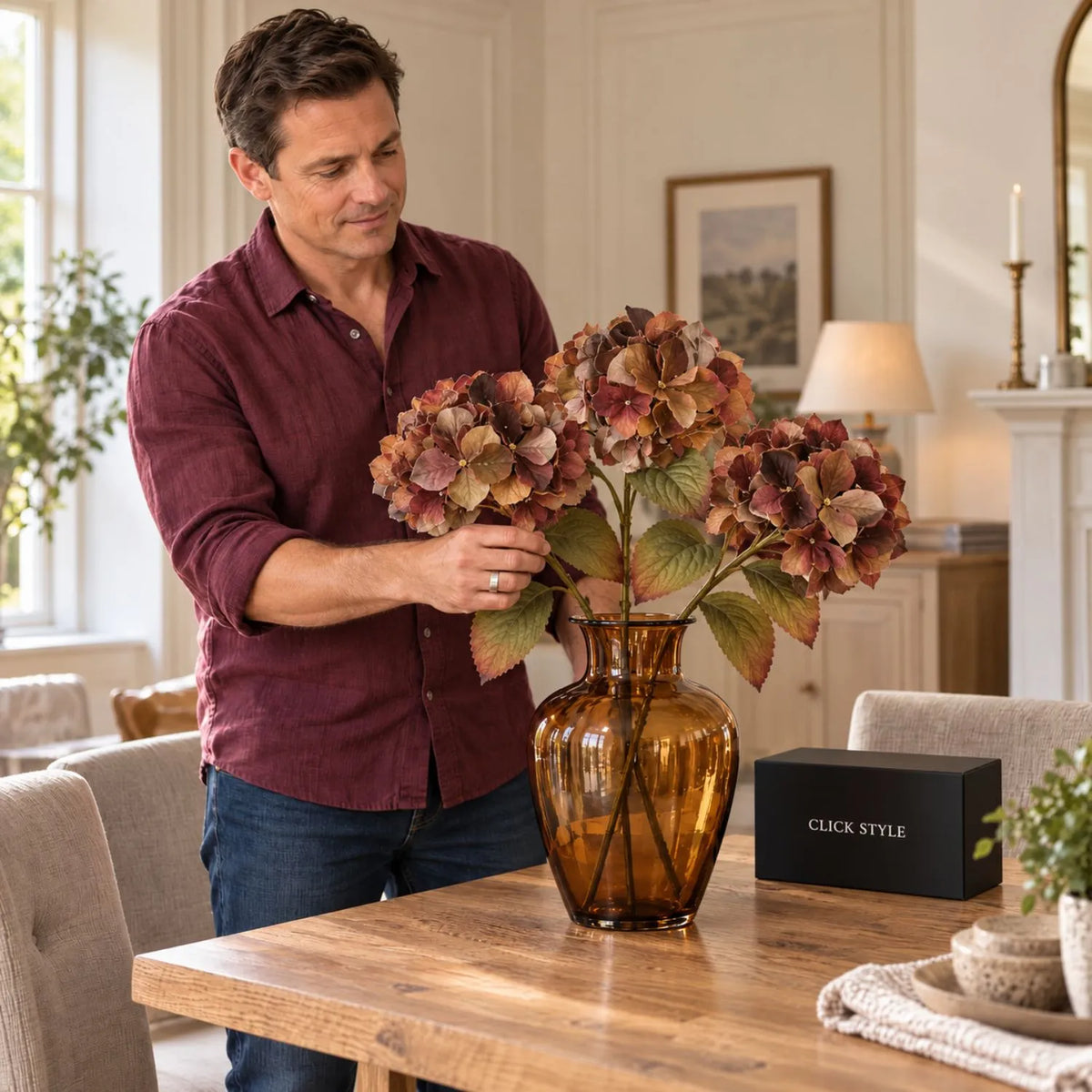 Artificial Russet Brown & Burgundy Hydrangea Stem with Realistic Autumn Foliage – Lifelike Faux Brown Hydrangea Flower 50x26x26cm – Man wearing a burgundy linen shirt arranging three autumn hydrangea stems in an amber glass vase on an oak dining table with black CLICK STYLE branding | Click Style