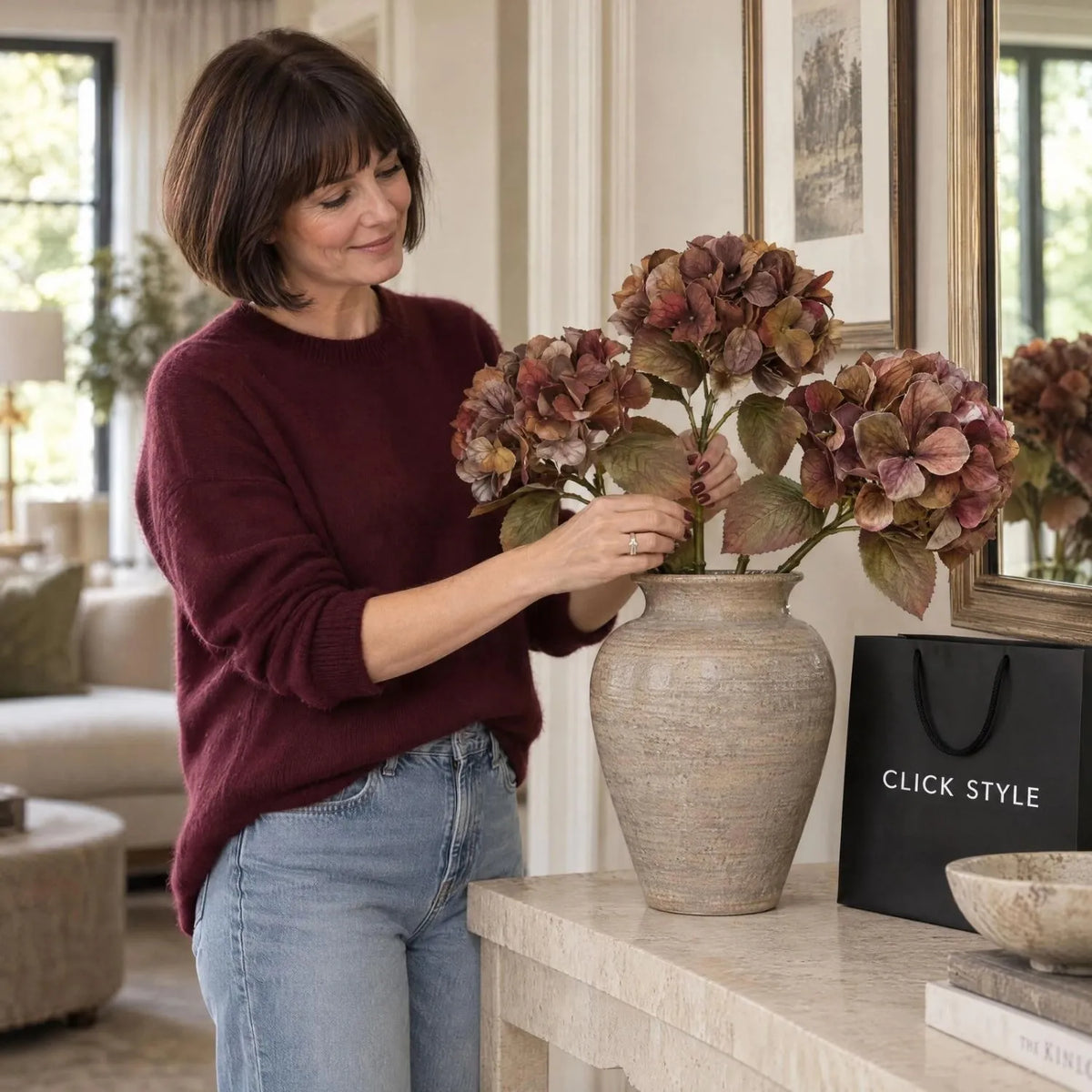 Artificial Russet Brown & Burgundy Hydrangea Stem with Realistic Autumn Foliage – Lifelike Faux Brown Hydrangea Flower 50x26x26cm – Woman wearing a burgundy cashmere jumper arranging three artificial hydrangea stems in a textured taupe vase beside a black CLICK STYLE shopping bag | Click Style