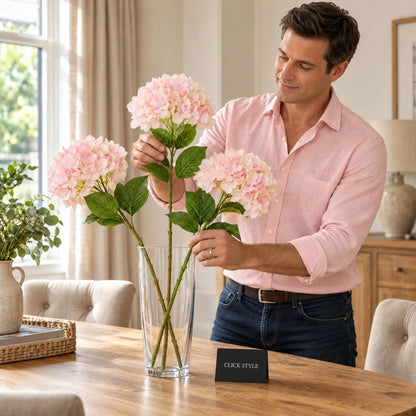 Artificial Soft Pink Long Stem Hydrangea with Full Rounded Flower Head & Realistic Green Leaves – Lifelike Pink Faux Hydrangea Flower 70cm displayed as three separate stems in a clear glass vase being arranged by a man wearing a pink shirt and dark blue jeans at a natural oak dining table | Click Style