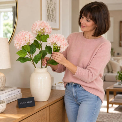 Artificial Soft Pink Long Stem Hydrangea with Full Rounded Flower Head & Realistic Green Leaves – Lifelike Pink Faux Hydrangea Flower 70cm shown as three individual stems being arranged in an ivory ceramic vase by a woman wearing a pink cashmere jumper and light blue jeans beside an oak sideboard | Click Style