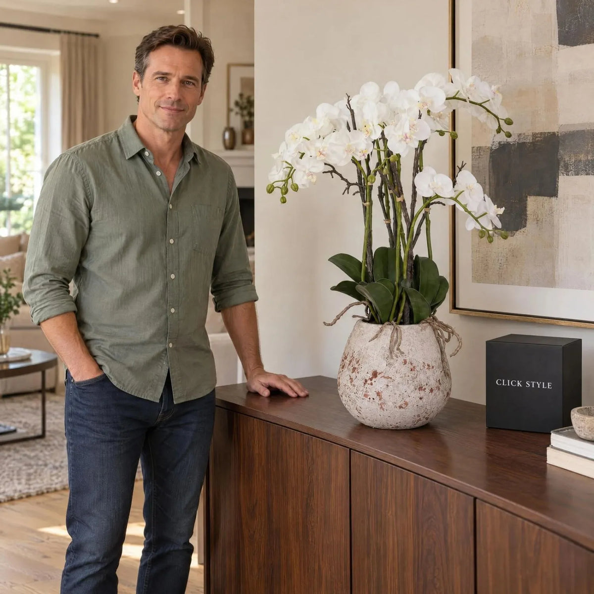 Artificial White Orchid with Cascading Blooms Dark Green Leaves & Realistic Roots in Weathered Stone Effect Pot – Large Potted Faux Orchid Arrangement 70x60x60cm styled on a dark walnut sideboard beside Damian with black CLICK STYLE presentation box | Click Style