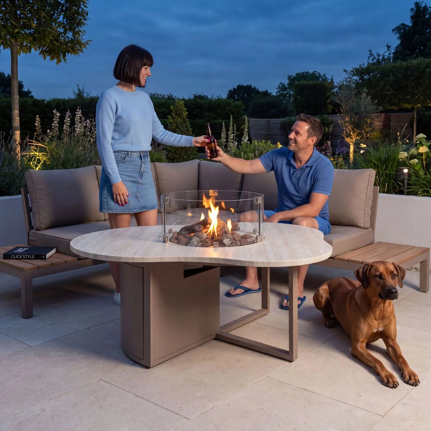 Cosiloft Novi Taupe Beige Organic Shaped Aluminium Outdoor Gas Fire Pit Table with Stone Look Top and Glass Wind Guard styled at dusk with a woman handing a seated man a beer and dog nearby 47x120x90cm | Click Style