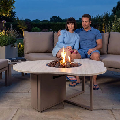 Cosiloft Novi Taupe Beige Organic Shaped Aluminium Outdoor Gas Fire Pit Table with Stone Look Top styled at dusk with a couple relaxing on an outdoor sofa 47x120x90cm | Click Style