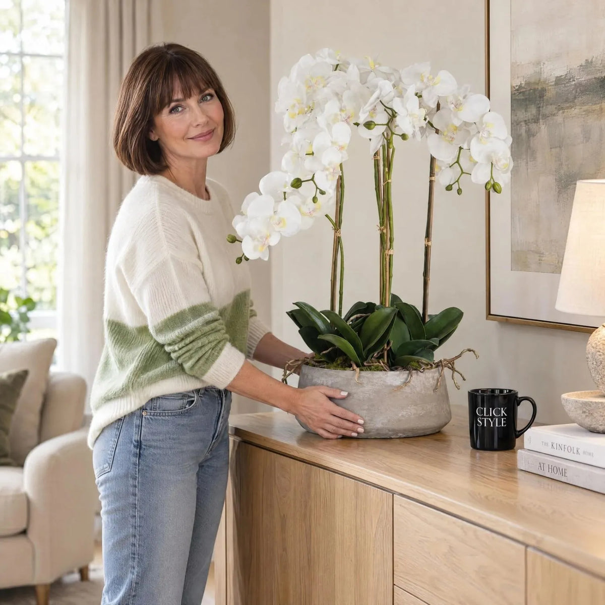 Large Artificial White Orchid in Grey Stone Effect Pot Being Positioned on an Oak Sideboard by a Woman in a White and Green Jumper | Click Style