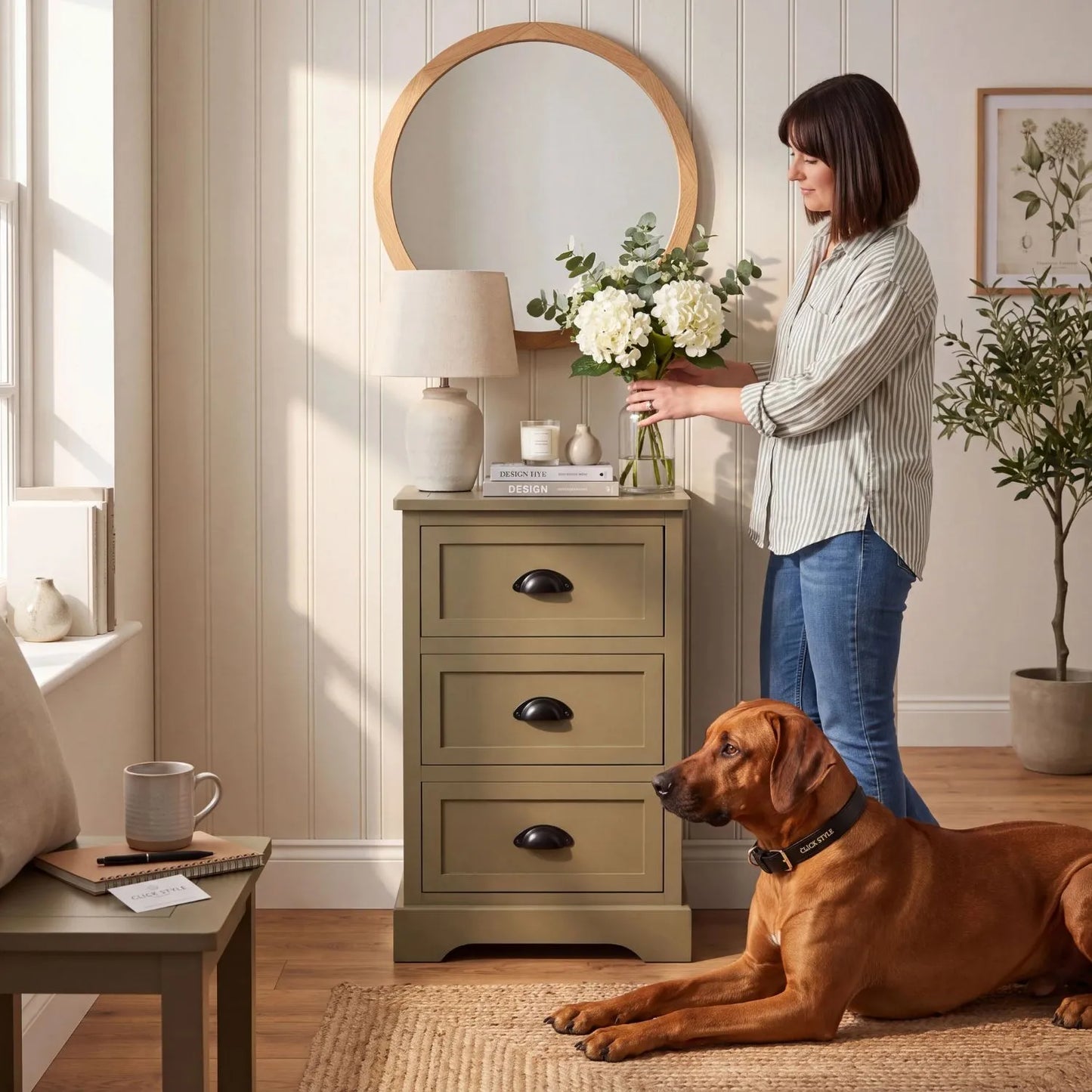Olive Green Pine Wood 3 Drawer Chest of Drawers with Matt Black Cup Handles Styled in a Modern Country Bedroom with Woman Arranging Flowers and Rhodesian Ridgeback Dog 70x41x33cm | Click Style