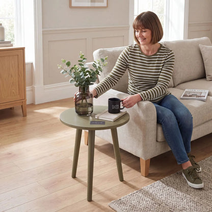 Olive Green Pine Wood Round Tripod Side Table styled in a modern country living room with a woman sitting on a beige sofa beside the table with a smoked glass vase and Click Style mug 42x40x40cm | Click Style