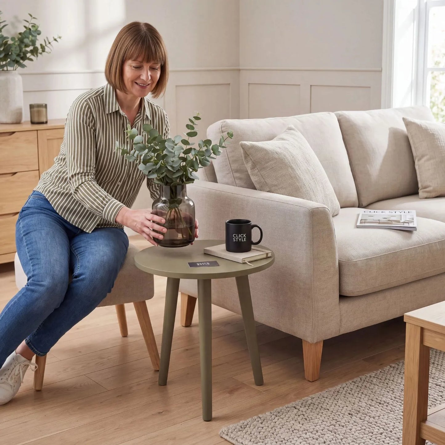 Olive Green Pine Wood Round Tripod Side Table styled in a modern country living room with a woman placing a smoked glass vase with eucalyptus onto the tabletop 42x40x40cm | Click Style