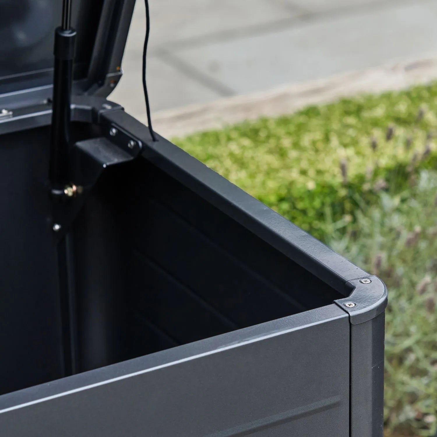 Small Black Galvanised Steel Outdoor Cushion Storage Box lifestyle image from front right with open lid showing gas piston and rounded front edge | Click Style