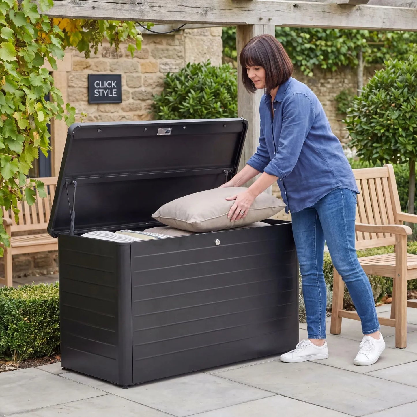 Small Black Galvanised Steel Outdoor Cushion Storage Box with Lockable Lid open with woman storing cushions in a British garden 78x134x62cm | Click Style