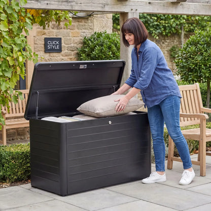 Small Black Galvanised Steel Outdoor Cushion Storage Box with Lockable Lid open with woman storing cushions in a British garden 78x134x62cm | Click Style