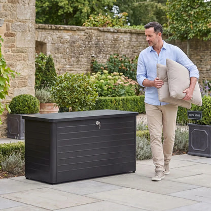 Small Black Galvanised Steel Outdoor Cushion Storage Box with Lockable Lid styled in a British garden with man carrying cushions 78x134x62cm | Click Style