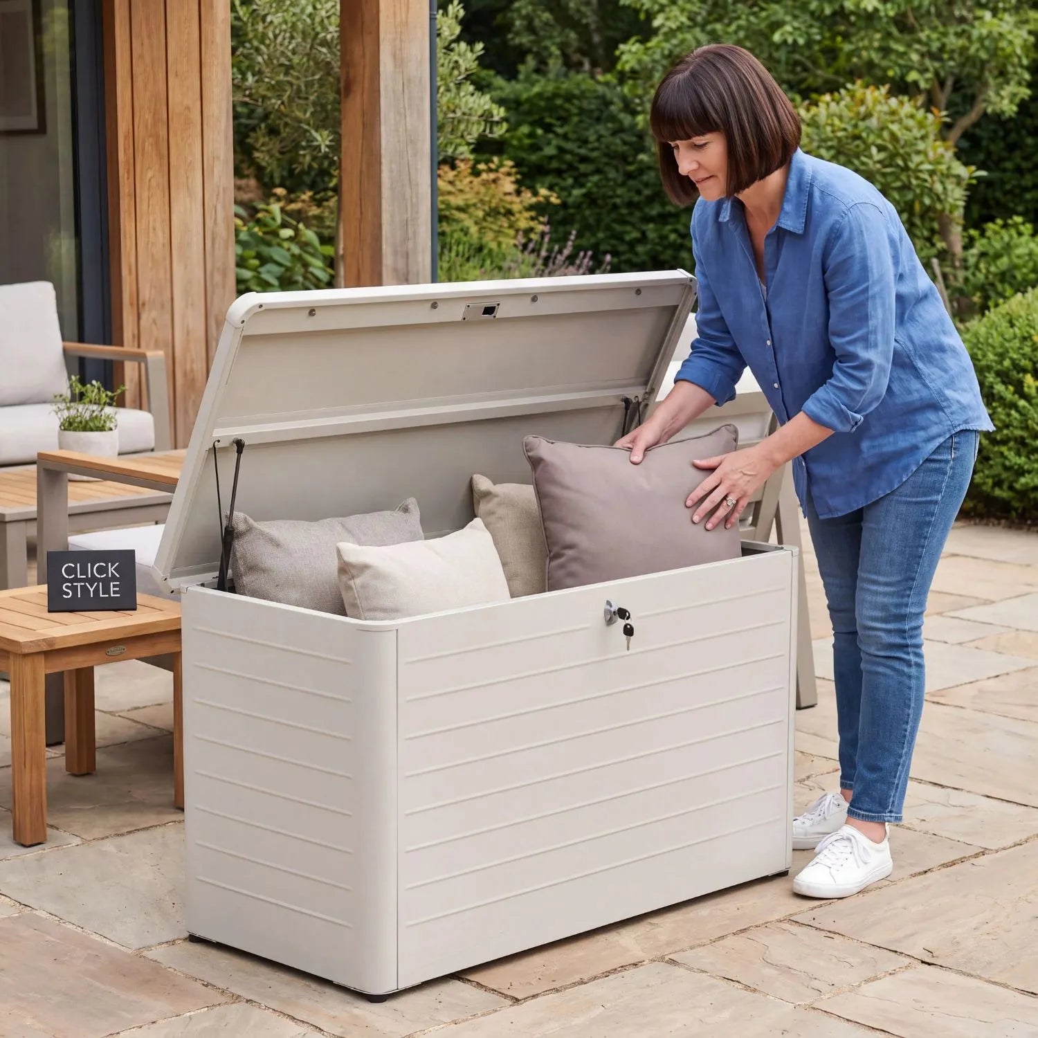 Small Light Grey Pebble Galvanised Steel Outdoor Cushion Storage Box with open lid and woman placing a garden cushion inside in a contemporary garden | Click Style