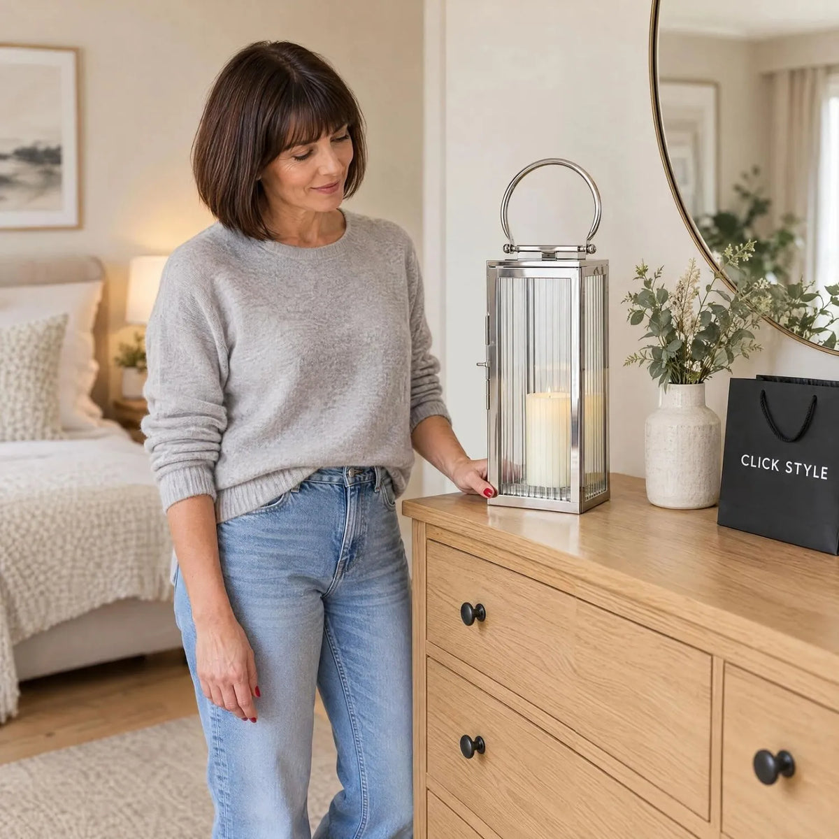 Small Polished Silver Nickel Candle Lantern with Ribbed Glass Panels & Curved Carry Handle – Decorative Stainless Steel Lantern 43x19x17cm being styled on an oak bedroom chest of drawers by a woman wearing a silver cashmere jumper and light blue jeans with red nails | Click Style