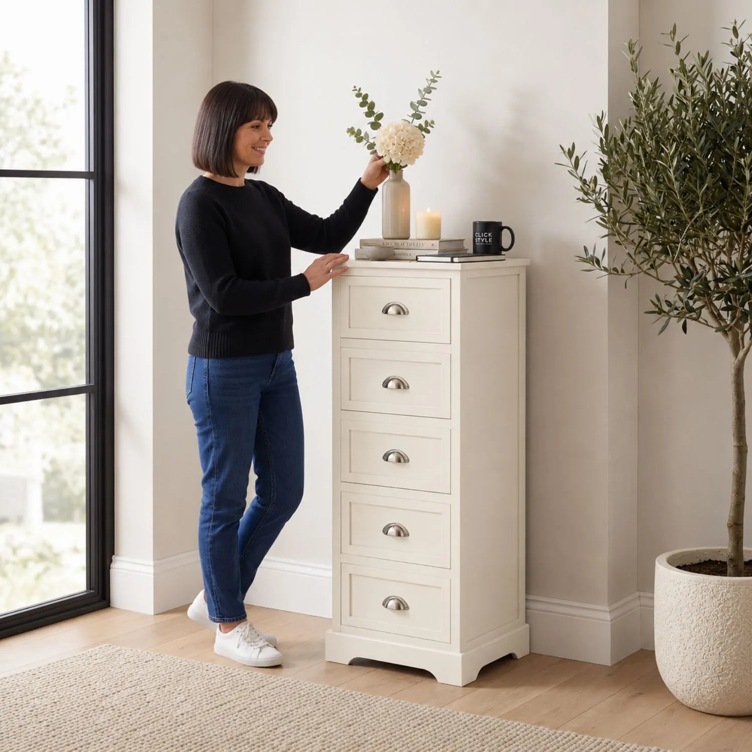 Warm Ivory Pine Wood 5 Drawer Tall Boy Chest of Drawers with Polished Silver Cup Handles styled in a modern home with a woman arranging flowers | Click Style