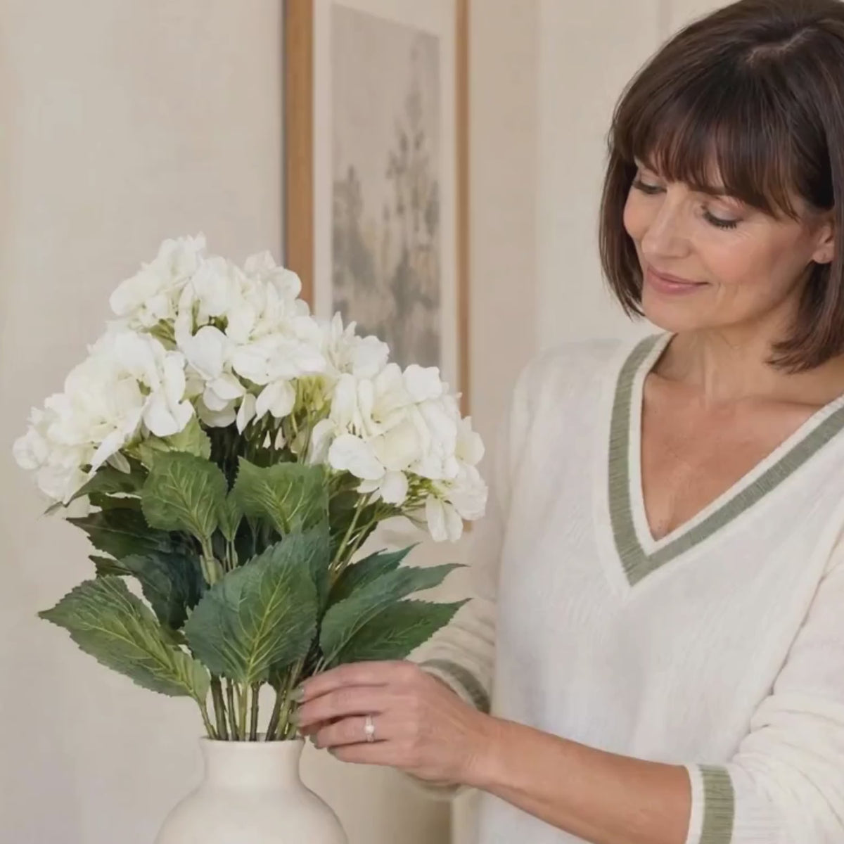 Artificial Creamy White Multi-Bloom Hydrangea Bouquet Stem with Realistic Green Foliage – Lifelike Faux Cream Hydrangea Arrangement 54x34x34cm – Lifestyle video showing a woman in a cream V-neck jumper with sage green trim gently arranging creamy white hydrangea blooms in a ceramic vase on an oak console table with CLICK STYLE branding | Click Style