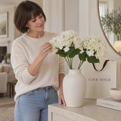 Artificial Creamy White Multi-Bloom Hydrangea Stem with Realistic Green Leaves – Lifelike Faux White Hydrangea Flower 47x23x23cm – Lifestyle video of a woman naturally adjusting creamy white artificial hydrangeas in an ivory ceramic vase beside a CLICK STYLE shopping bag | Click Style