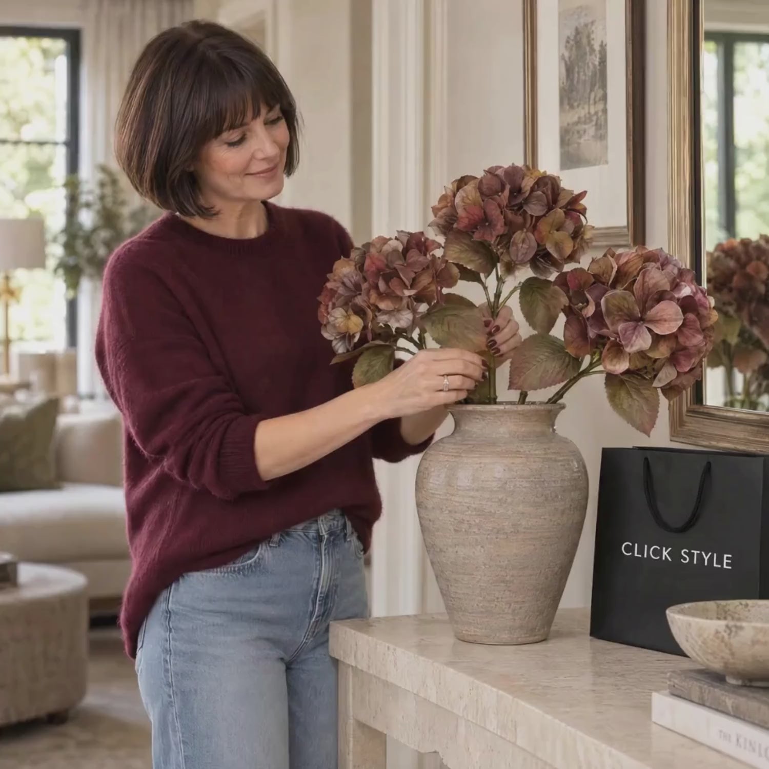 Artificial Russet Brown & Burgundy Hydrangea Stem with Realistic Autumn Foliage – Lifelike Faux Brown Hydrangea Flower 50x26x26cm – Lifestyle video showing a woman in a burgundy cashmere jumper naturally arranging three autumn hydrangea stems in a textured taupe vase beside a black CLICK STYLE shopping bag | Click Style