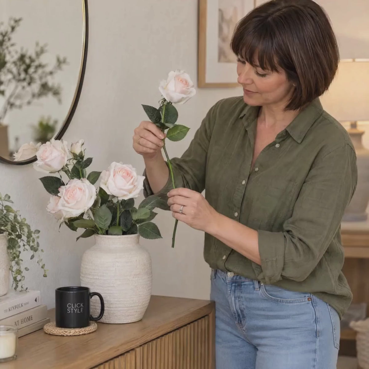 Artificial Pale Pink Tea Rose Stem with Realistic Green Foliage – Lifelike Pink Faux Rose Flower 65cm – Lifestyle video showing a woman in an olive green shirt gently holding a pale pink tea rose stem beside a textured cream vase filled with pink roses on an oak sideboard with a black CLICK STYLE branded mug | Click Style