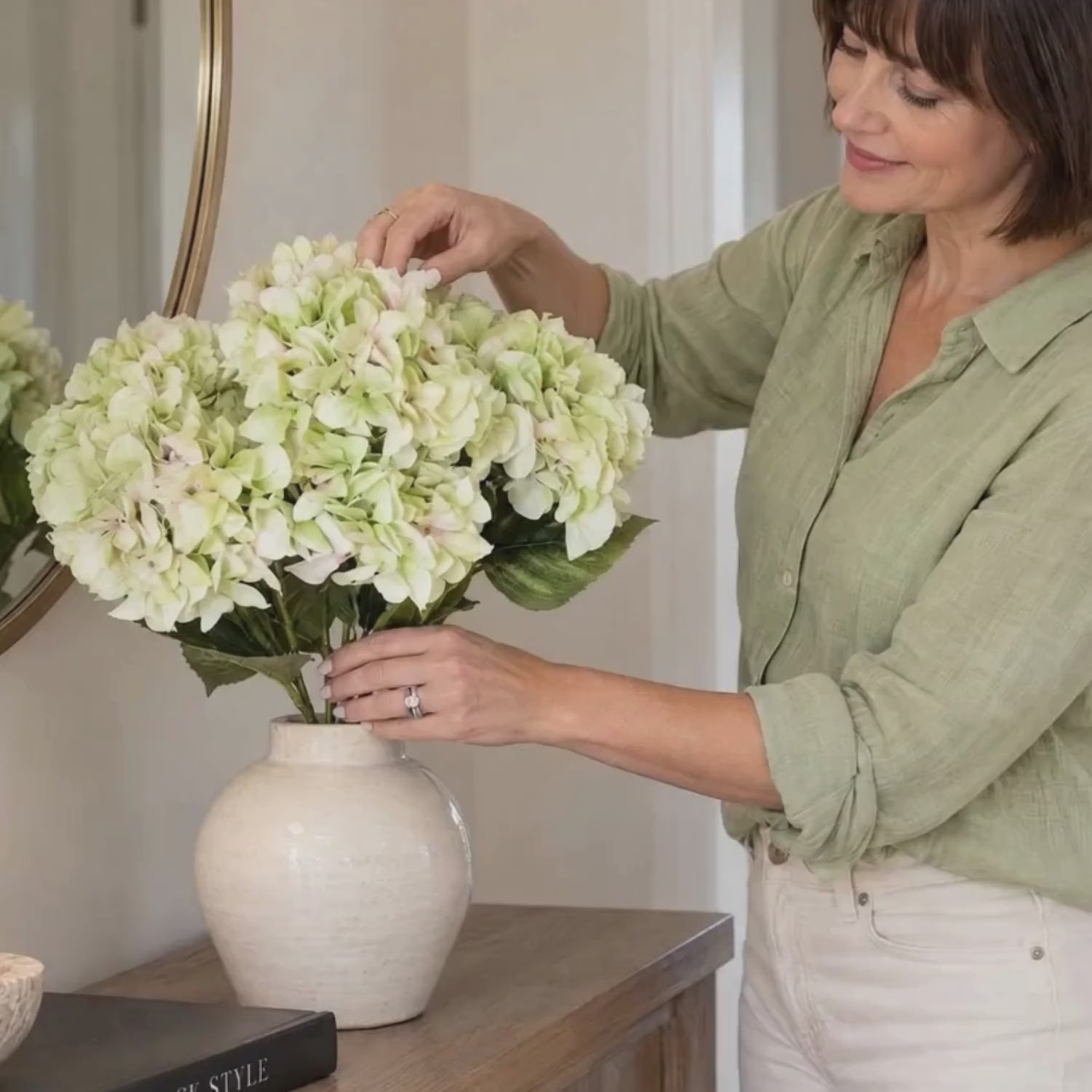 Artificial Pale Green Multi-Bloom Hydrangea Bouquet Stem with Realistic Foliage – Large Lifelike Faux Light Lime Green Hydrangea 58cm – Lifestyle video showing a woman in a sage green linen shirt gently arranging pale green hydrangea flowers in a cream ceramic vase on an oak console table with CLICK STYLE branding | Click Style