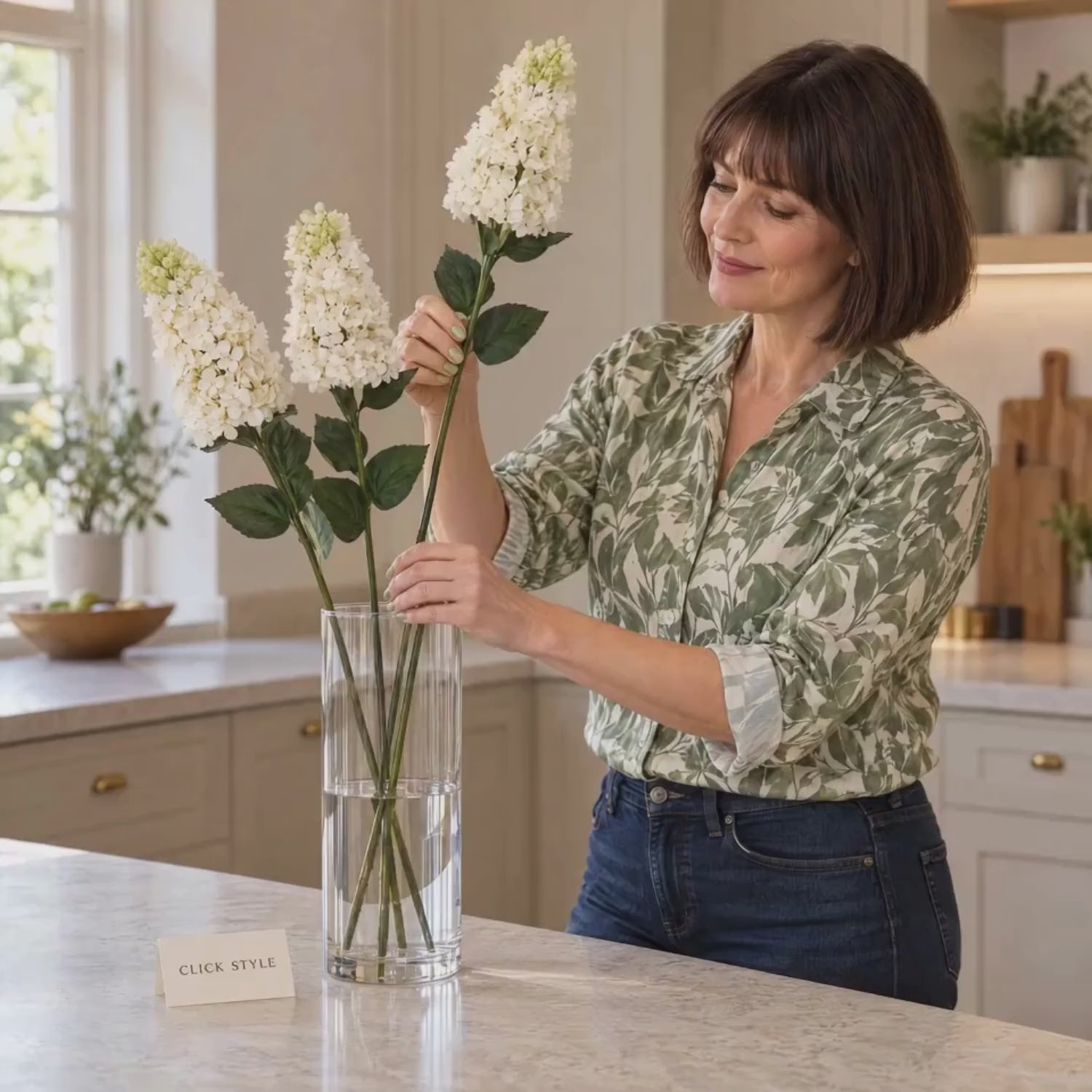 Artificial Creamy White Long Stem Spear Hydrangea with Pointed Flower Head & Realistic Green Leaves – Tall Faux Cream Hydrangea Flower 88cm – Lifestyle video showing a woman in a sage green patterned shirt gently arranging three creamy white spear hydrangea stems in a clear glass vase on a kitchen worktop with CLICK STYLE branding | Click Style