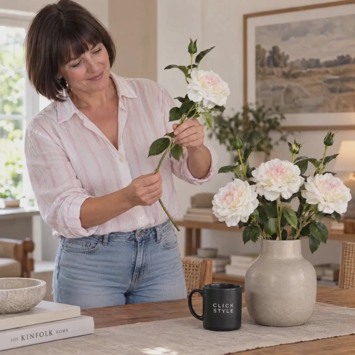 Artificial White Garden Rose Spray with Open Bloom & Rosebud – Lifelike White Faux Rose Flower with Pale Pink Centre & Realistic Green Foliage 68cm – Lifestyle video showing a woman in a pale pink striped shirt gently holding a white garden rose spray with blush pink centre and green rosebud beside a ceramic vase of roses on a wooden table with a black CLICK STYLE branded mug | Click Style