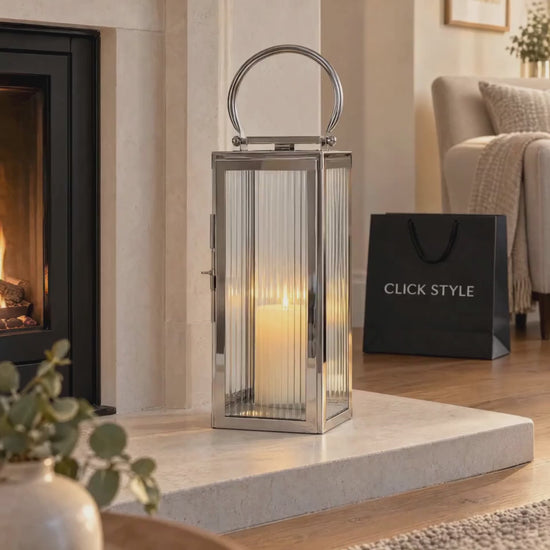 Small Polished Silver Nickel Candle Lantern with Ribbed Glass Panels & Curved Carry Handle – Decorative Stainless Steel Lantern 43x19x17cm with gently flickering candlelight on a limestone fireplace hearth in a neutral living room with black CLICK STYLE branding | Click Style