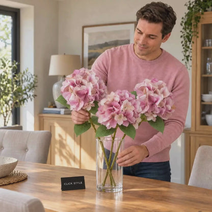 Artificial Pink Hydrangea Stem with Multi-Tonal Petals & Realistic Green Leaves – Lifelike Faux Pink Hydrangea Flower 50x30x30cm shown in a lifestyle video of a man wearing a pink jumper gently arranging three individual hydrangea stems in a clear glass vase on an oak dining table, displaying realistic pink, mauve and cream flower heads with serrated green leaves | Click Style