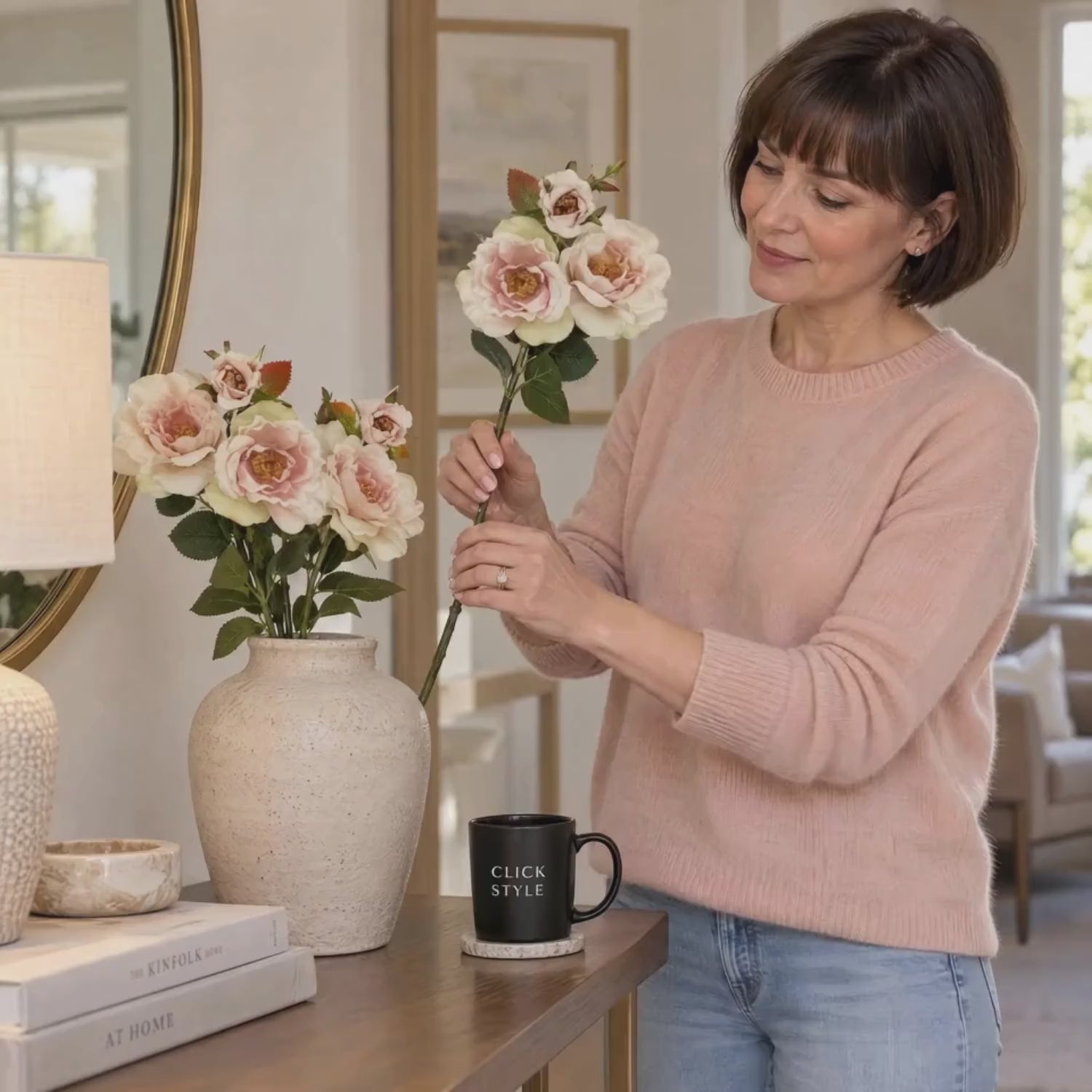 Artificial Pale Blush Tea Rose Stem with Open Blooms & Rosebud – Lifelike Palest Blush Pink Faux Rose Flowers with Realistic Green Foliage 60cm – Woman wearing a peach cashmere jumper gently arranging the pale blush tea rose stem beside matching artificial roses in a cream ceramic vase on an oak console table | Click Style